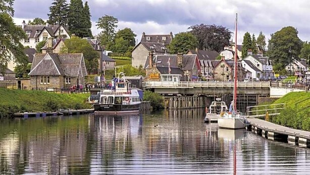 A calm river with boats, a dock, and houses in the background. - Loch Ness Gate House Apartments (Fort Augustus)