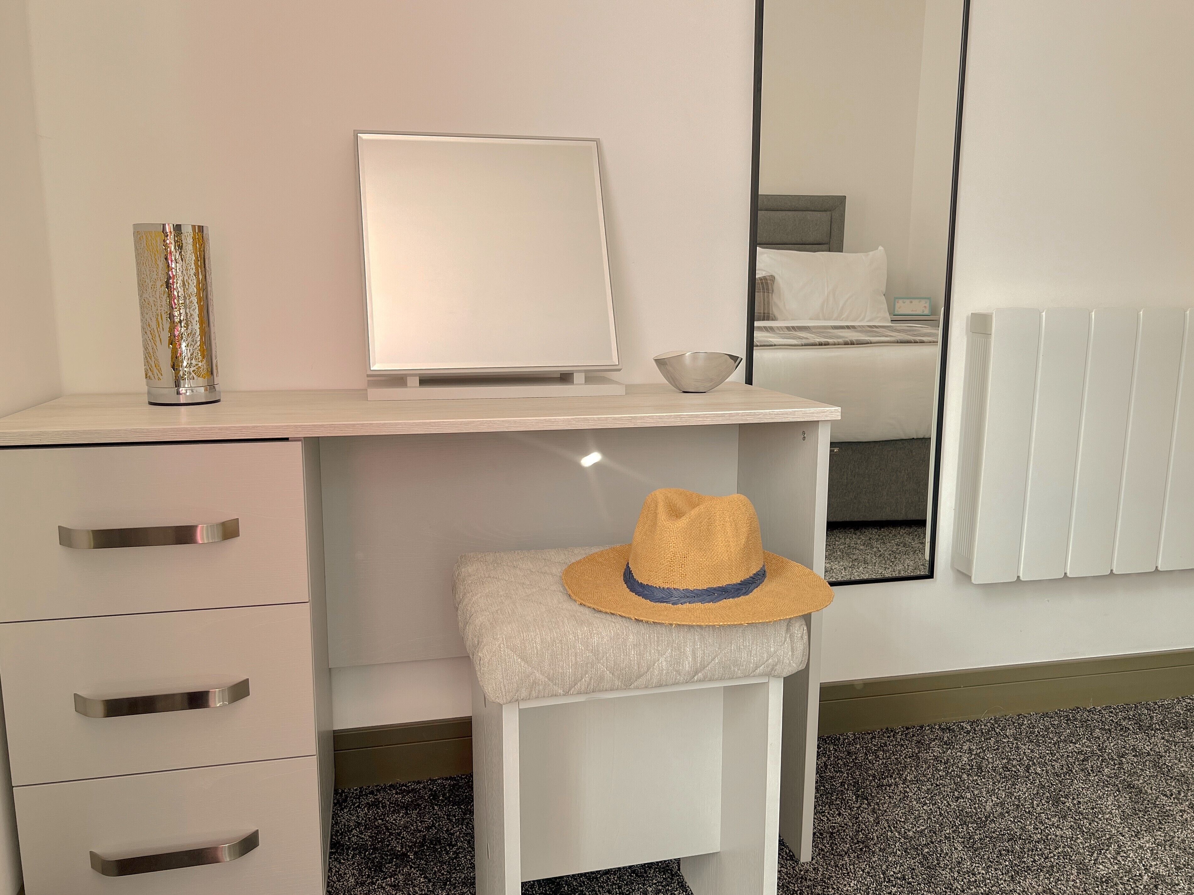 Habitación de hotel con un escritorio blanco, un espejo, un taburete pequeño con un sombrero y un radiador.