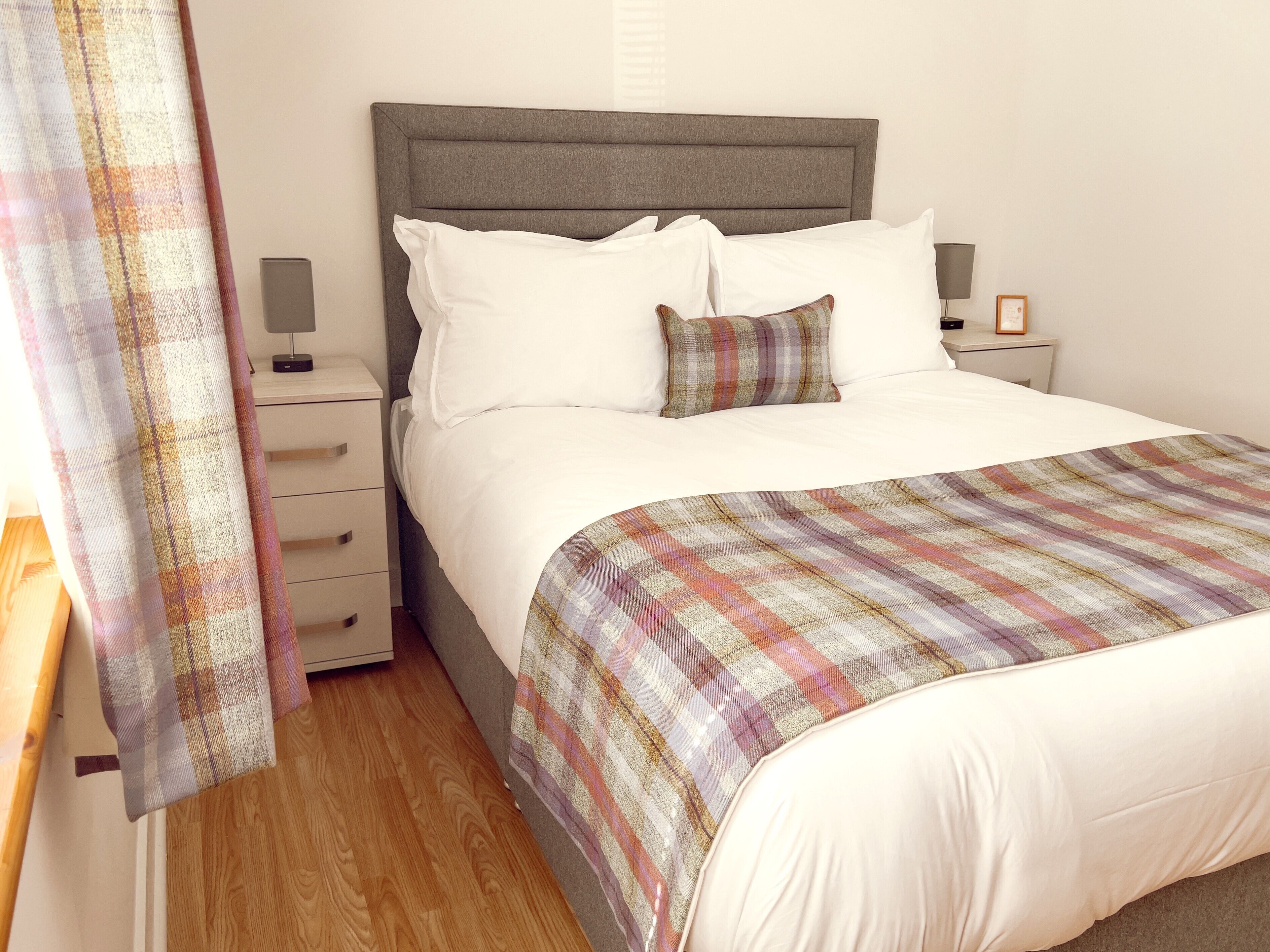 Een net opgemaakt bed met een geruit plaid, een grijs gestoffeerd hoofdbord en witte beddengoed. Er staat een nachtkastje met een lamp en een ingelijste foto.