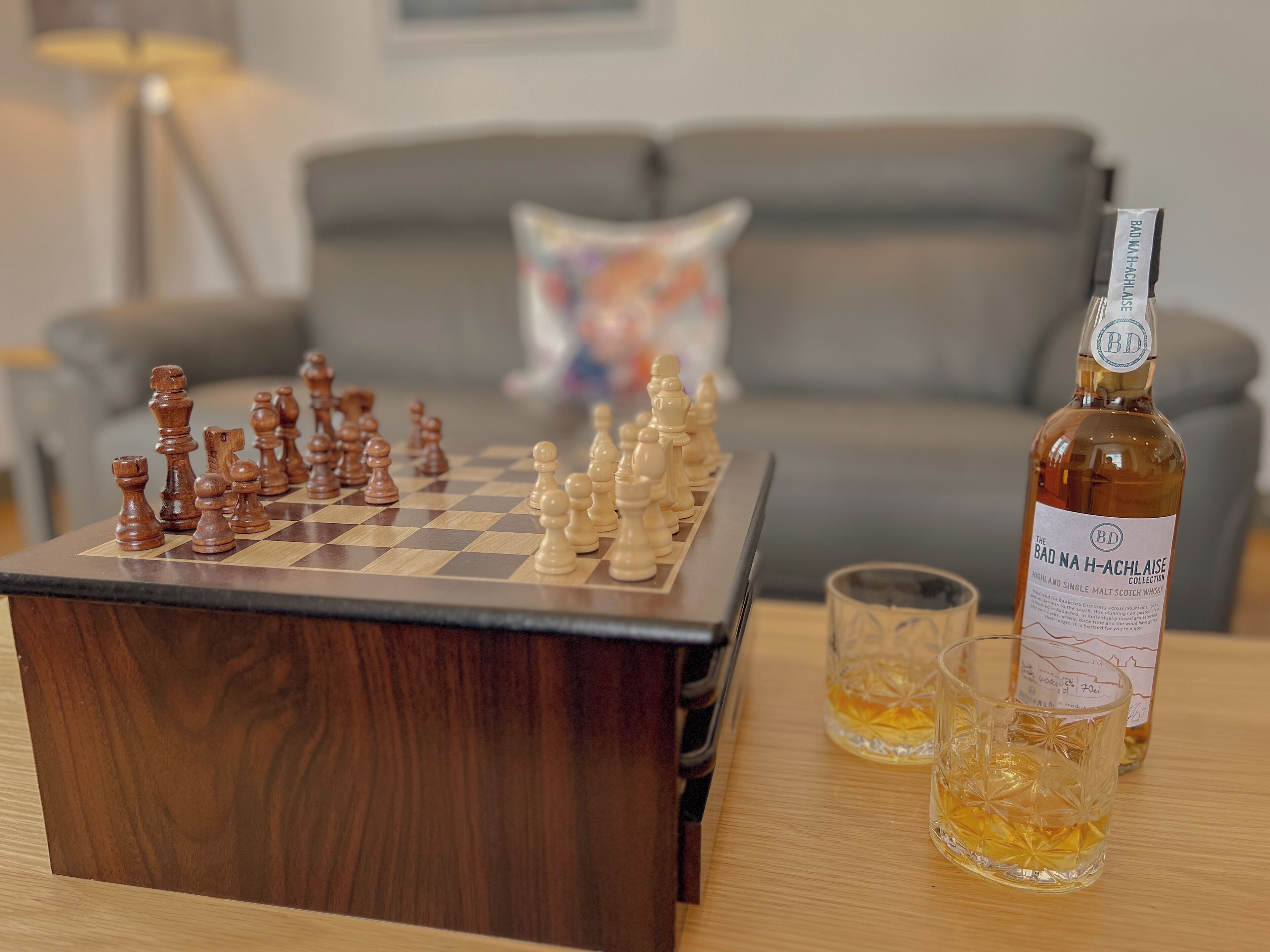A wooden chess table with chess pieces, a bottle of whisky, and two glasses on a wooden table.