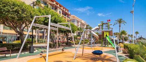 A playground with swings and slides, surrounded by greenery and buildings.