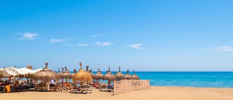 A beach with thatched umbrellas and lounge chairs, a clear blue sky, and calm sea.