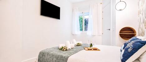 Une chambre à coucher comprenant un lit, un téléviseur à écran plat, une fenêtre avec des rideaux, une porte en bois et un luminaire fixé au mur.