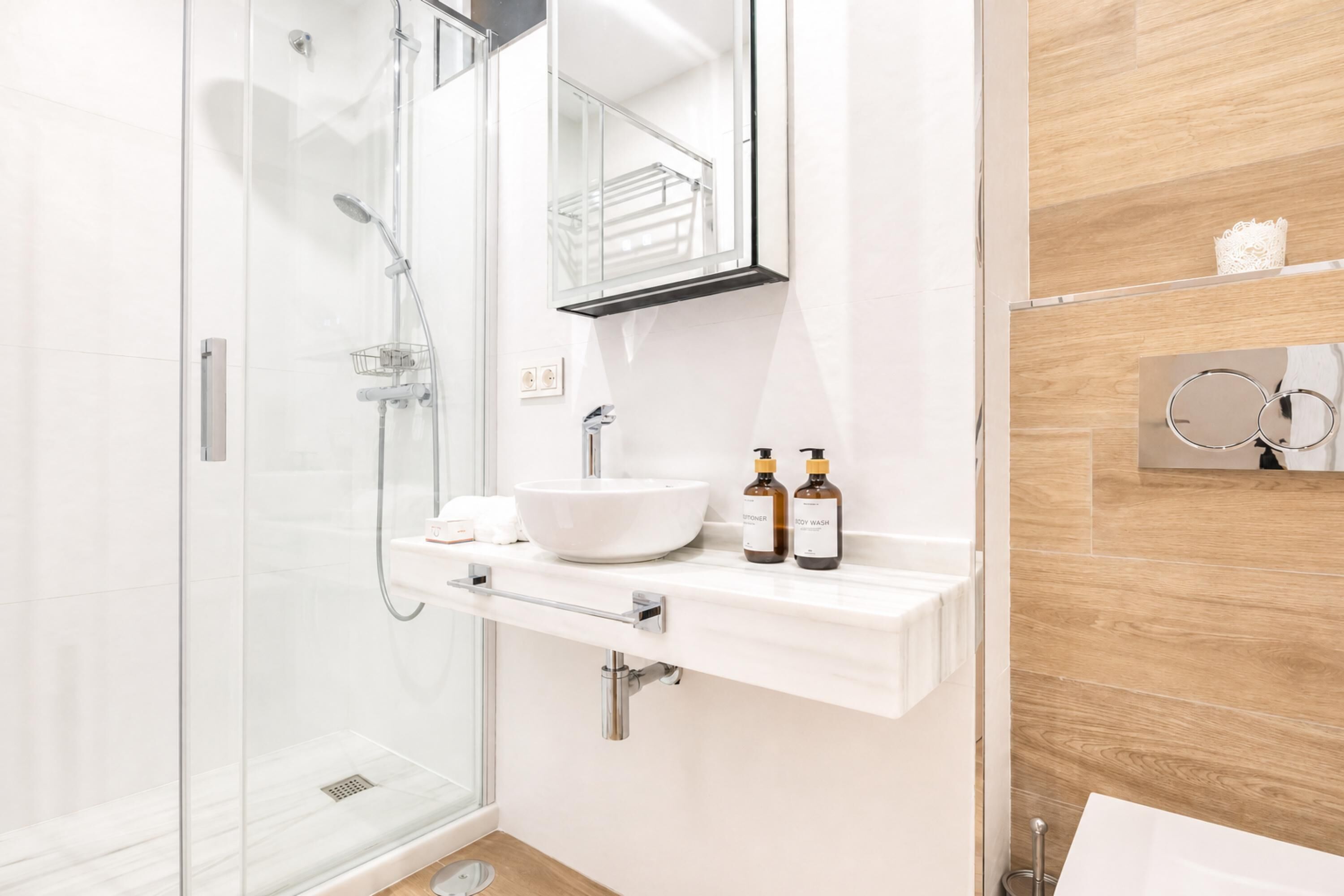 A modern bathroom with a glass shower enclosure, a white sink, and a vanity with a mirror. There are two bottles of lotion on the vanity.