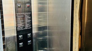 An elevator interior with a Titan Elevators sign, emergency contact numbers, and a maximum capacity sign. - Maharishi Inn (Faizabad)