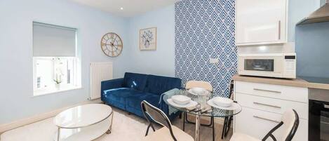A compact living space with a kitchenette, a blue sofa, a dining table with chairs, and a small round table with plates.