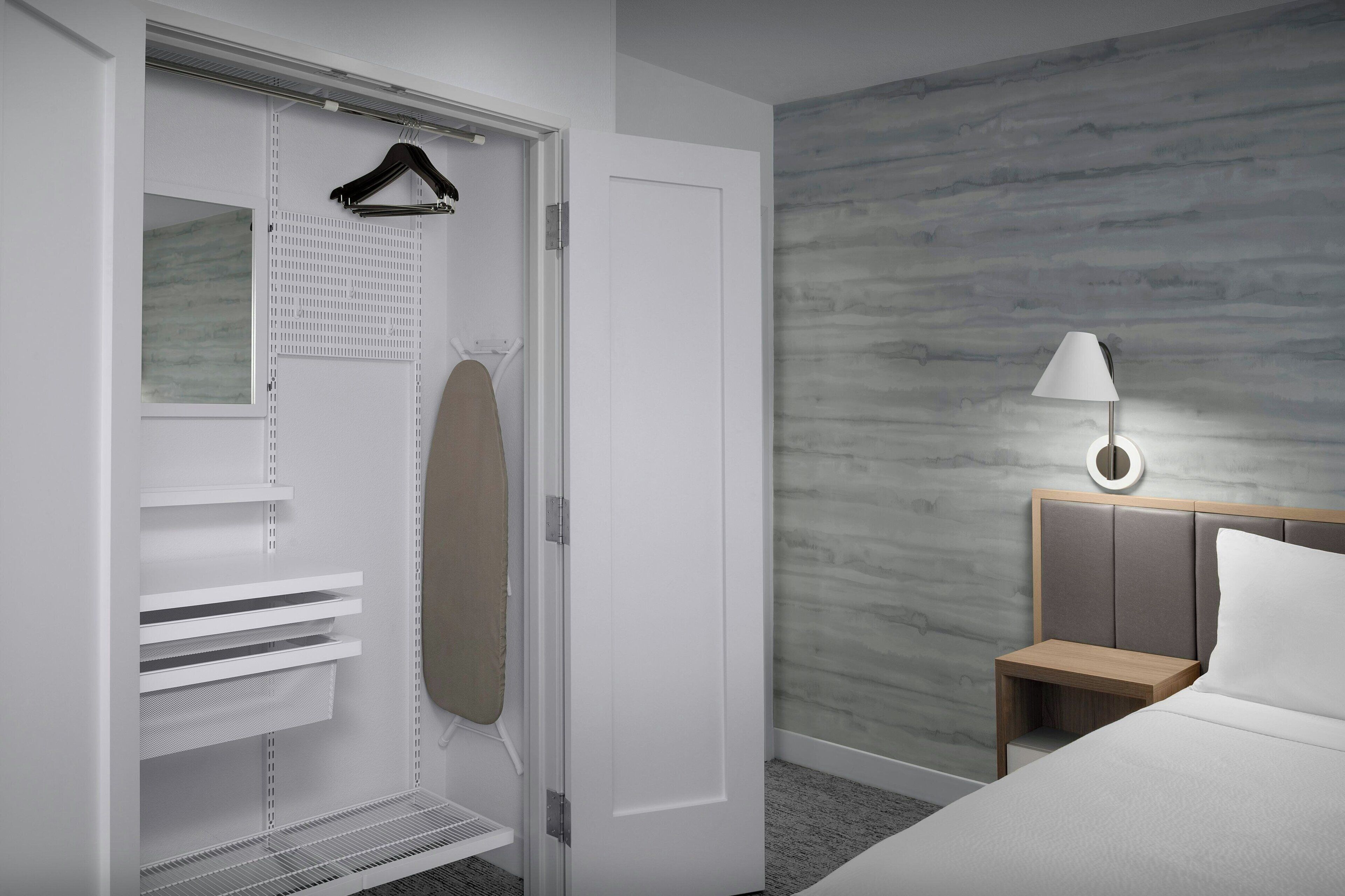 A hotel room with a walk-in closet, a bed with a wooden headboard, a bedside table with a lamp, and a wall with a textured pattern.