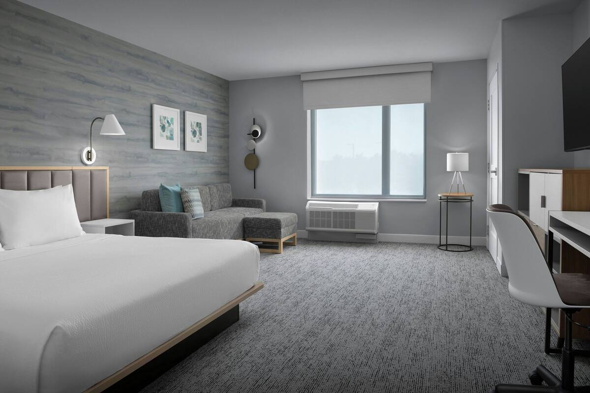 Una habitación de hotel moderna con una cama grande, un sofá, un escritorio y una silla. Se ve una ventana con vistas a la ciudad.