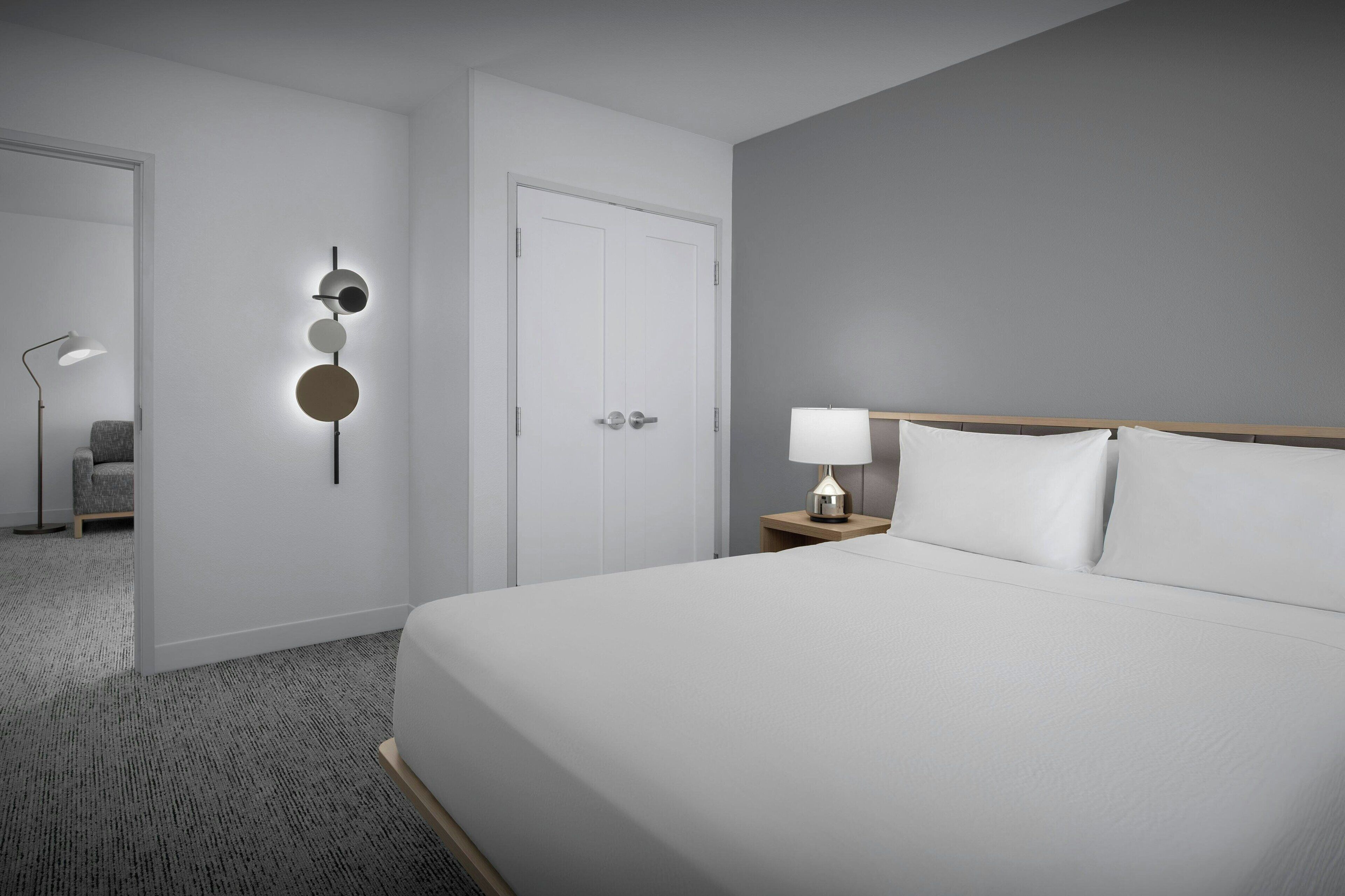 Camera d'albergo con un letto grande, un comodino con una lampada, un punto luce a parete e una porta.