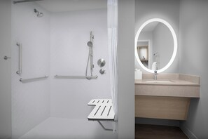 Ein modernes Badezimmer mit Dusche, Haltegriffen, einem Waschtisch inklusive Spiegel und einer Toilette mit Sicherheitsbügel.