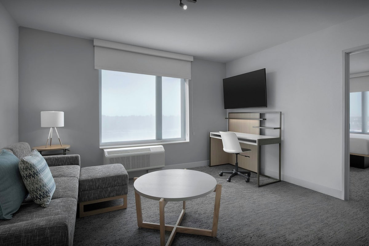 Una habitación de hotel moderna con un sofá gris, una mesa redonda, un escritorio con una silla, un televisor de pantalla plana y un ventanal con persianas.