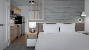 Una habitación de hotel moderna con cama, mesitas de noche, lámpara, alacenas y una cocina integrada con refrigerador, microondas y armarios.