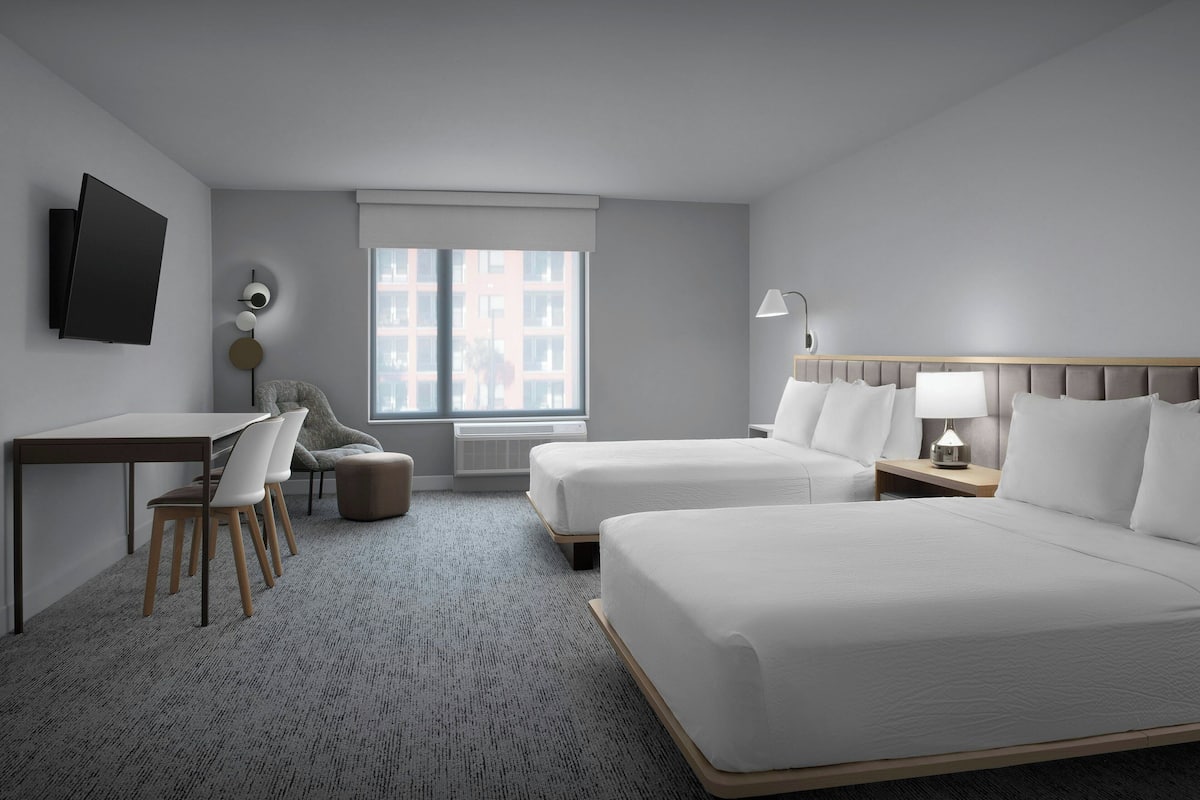 Habitación de hotel con dos camas, un escritorio, sillas, un televisor y una ventana con vista a edificios.