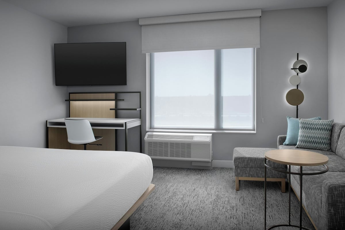 Una habitación de hotel moderna con cama, escritorio, silla, sofá, mesa redonda y un amplio ventanal con vistas.