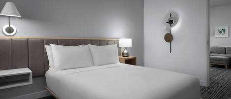 A modern hotel room with a large bed, a bedside table with a lamp, a wall clock, and a small shelf.