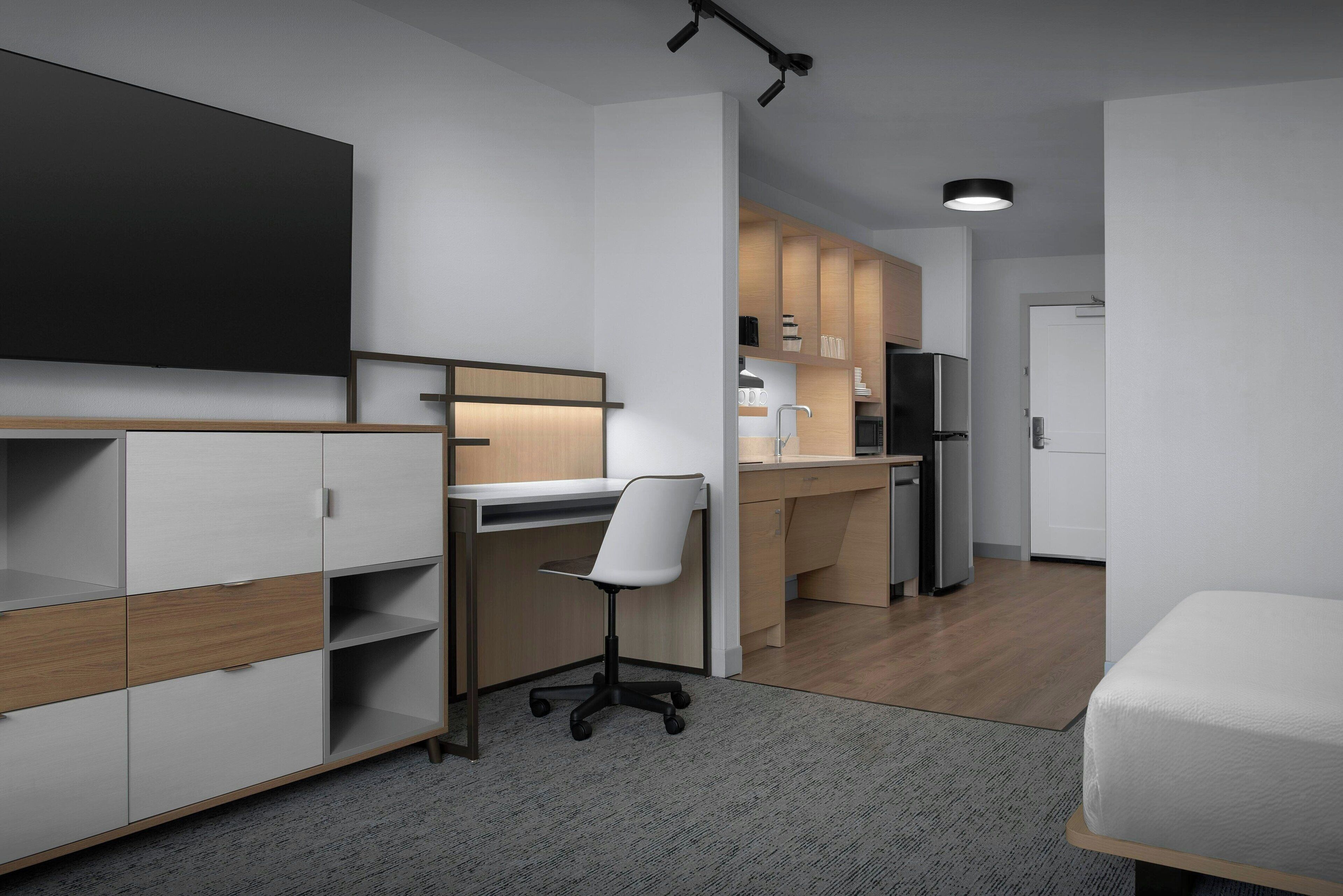 A modern, compact room with a desk, chair, kitchenette, and a bed.