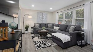 Ein modernes Wohnzimmer mit einem grauen Ecksofa, einem runden Couchtisch und einem großen Fenster mit weißen Vorhängen.