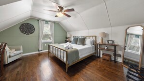 A bedroom with a sloped ceiling, a bed, a nightstand, a chair, a mirror, and a window with curtains. - Great Home_Roomy_Sleeps 13_Mins To GEHA_Dogs OK (Lee's Summit)