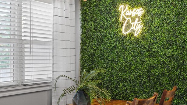 A dining area with a wooden table and chairs, a potted plant, and a neon sign on a green wall. - Great Home_Roomy_Sleeps 13_Mins To GEHA_Dogs OK (Lee's Summit)