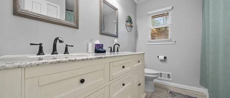 Un baño con tocador doble lavabo, inodoro, ventana con persianas y cortina de ducha.
