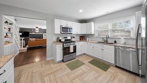 A modern kitchen with white cabinets, stainless steel appliances, and a herringbone wood floor. - Great Home_Roomy_Sleeps 13_Mins To GEHA_Dogs OK (Lee's Summit)