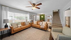 A living room with a brown leather sofa, a small table with a lamp, a ceiling fan, and a staircase. - Great Home_Roomy_Sleeps 13_Mins To GEHA_Dogs OK (Lee's Summit)