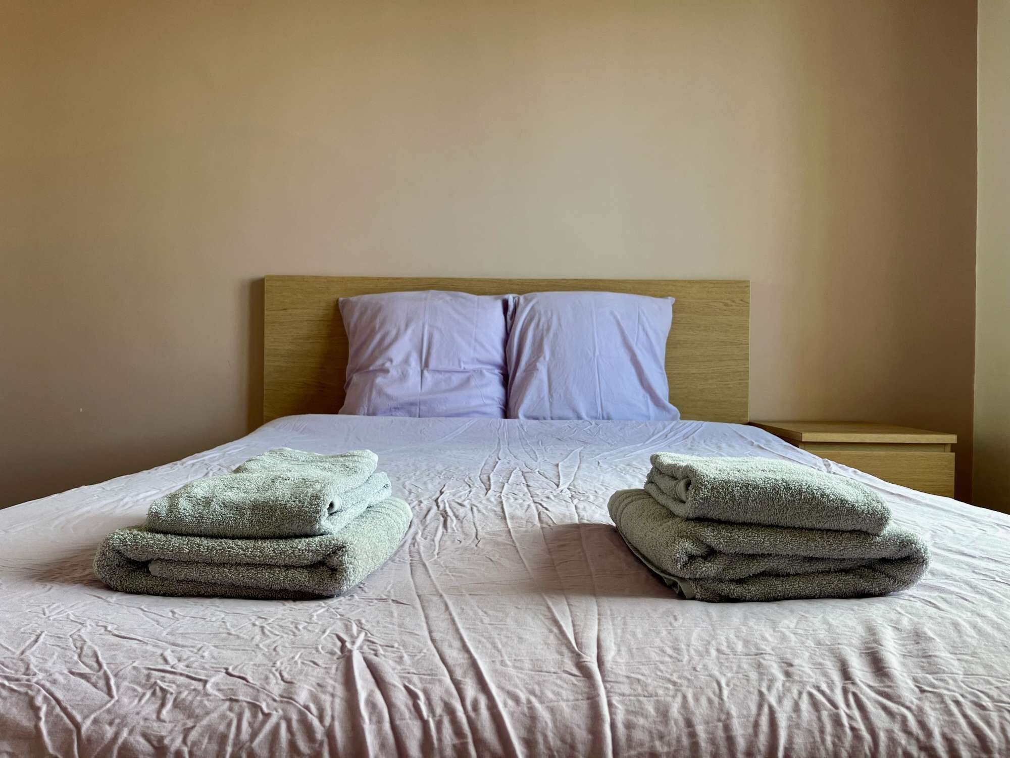 A neatly made bed with a wooden headboard, two pillows, and two stacks of folded towels.