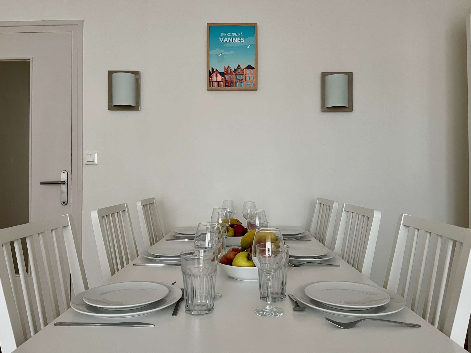 A dining table set for six with white chairs, a framed picture above, and two wall sconces.