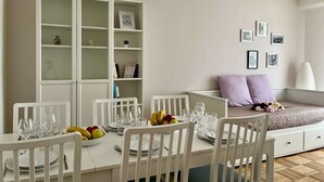 A dining area with a white table set for four, surrounded by white chairs. A white cabinet with glass doors is to the left, and a white sofa with a cushion is to the right.