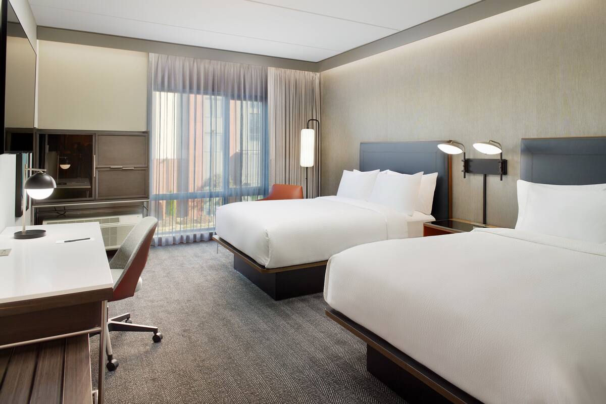 Ein Hotelzimmer mit zwei Betten, einem Schreibtisch, einem Stuhl und einem großen Fenster mit Vorhängen.