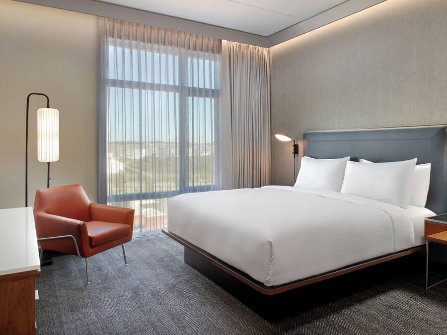 A hotel room with a large bed, a desk, a chair, a nightstand, and a lamp.