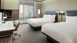Ein Hotelzimmer mit zwei Betten, einem Schreibtisch, einem Stuhl, einem Fernseher und einem großen Fenster mit Vorhängen.