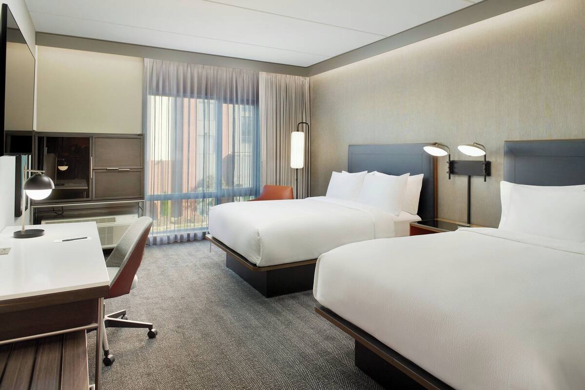 Ein Hotelzimmer mit zwei Betten, einem Schreibtisch, einem Stuhl, einem Fernseher und einem großen Fenster mit Vorhängen.