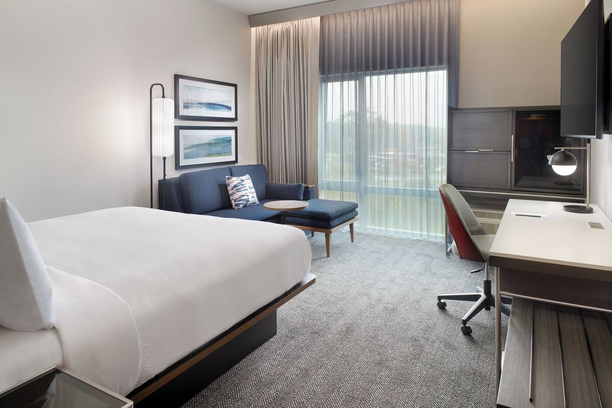 Ein Hotelzimmer mit Bett, Schreibtisch, Stuhl, Fernseher und einem fensterbepflanzten Fenster.