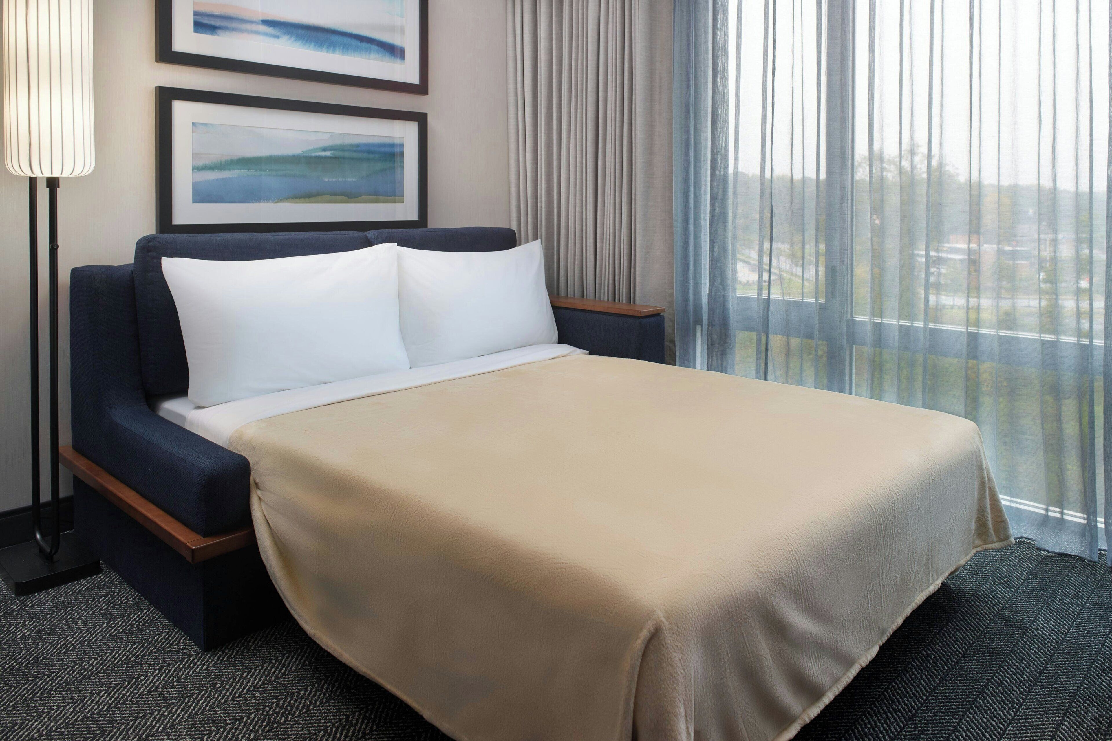 Una habitación de hotel con cama, mesita de noche, lámpara y ventana con cortinas.