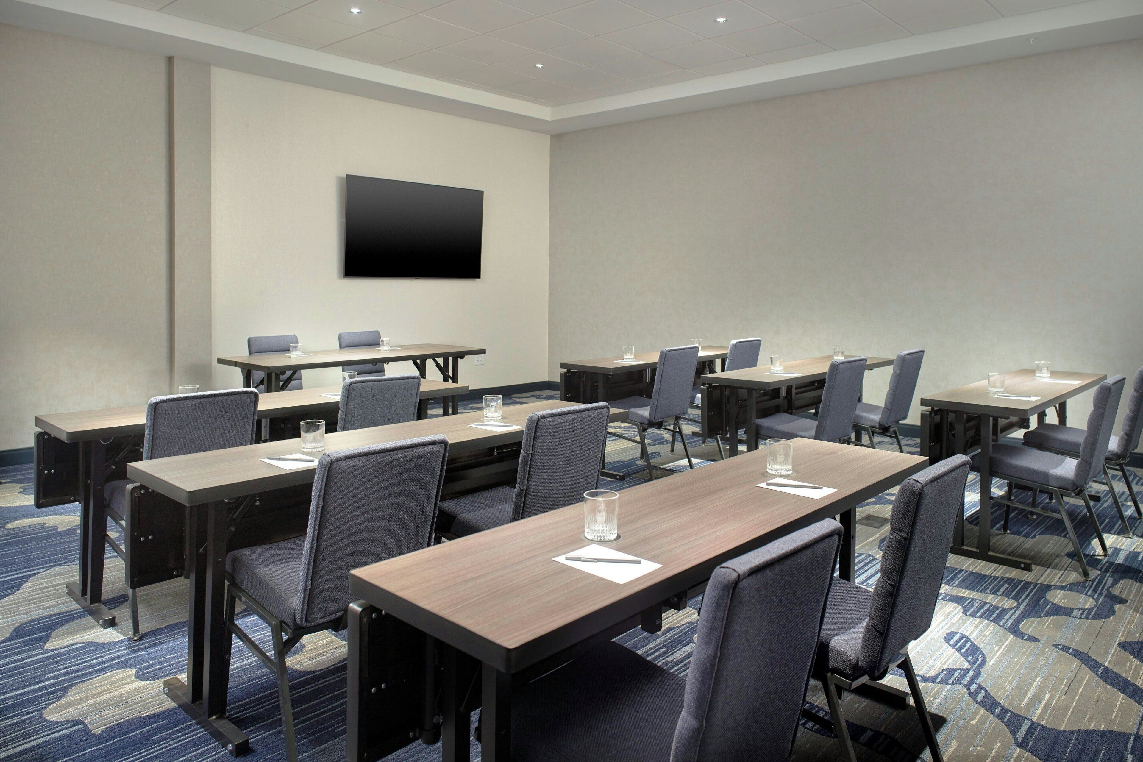 A conference room with multiple tables and chairs, a flat-screen TV mounted on the wall, and a carpeted floor.