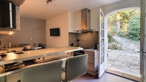 A modern kitchen with a built-in oven, a dining area with a table and chairs, and a view of a stone pathway and greenery outside. - Exclusive Secluded Villa: Truffle Oak Forest, Private Pool & Panoramic Lake View (Lissac sur Couze)