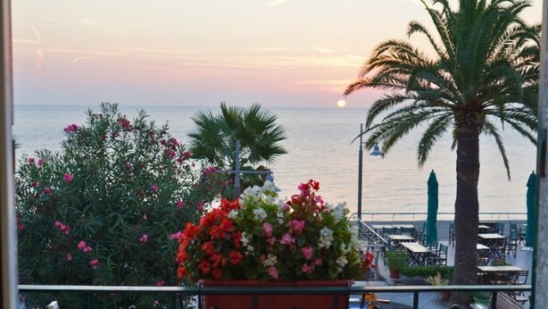 Un balcone con un tavolo apparecchiato per due, con vista su un tramonto sul mare, sullo sfondo palme e fiori.