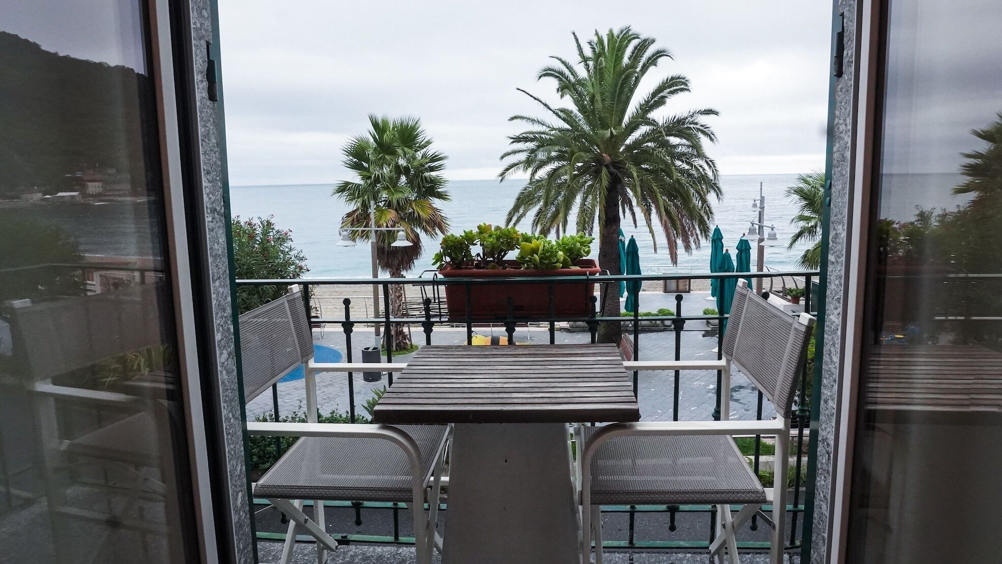 A balcony with two chairs and a table, overlooking a beach with palm trees and a view of the sea.