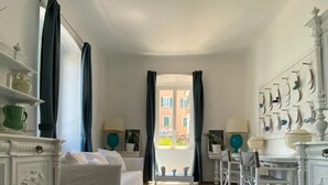 Una camera d'albergo con un letto, un tavolo da pranzo, sedie, un piccolo tavolo con una lampada, una mensola a parete con oggetti decorativi e vista sull'esterno.