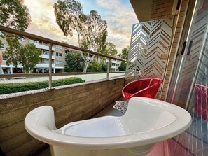 A balcony with a white bathtub, a red chair, and a view of a building with a chevron-patterned wall. - 🌿 Central Subiaco Oasis • 1BR • 1PK!<br>🏡 Peaceful inner-city retreat<br>🌳 Private balcony • Mueller Park<br>🍳 Fully equipped kitchen<br>🚆 Station 3-min walk<br>🚗 Free gated parking<br>🧺 Shared laundry<br>🪜 1st floor • 15 stairs<br>💗 Love Wh (Subiaco)