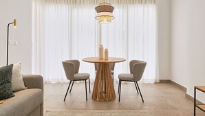A modern living room with a round wooden table, two grey chairs, a sofa with cushions, a floor lamp, and a hanging light fixture. - Solaga - Soho Suites (Málaga)
