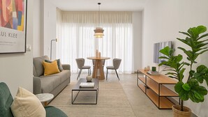 A modern living room with a sofa, coffee table, and a dining area with chairs and a table. - Solaga - Soho Suites (Málaga)