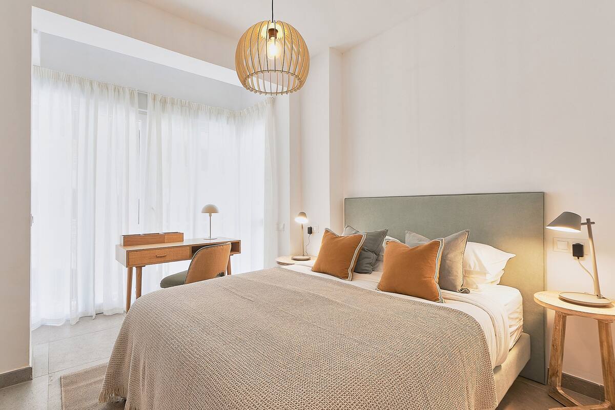 A hotel room with a bed, bedside tables, a desk, and a large window with curtains.