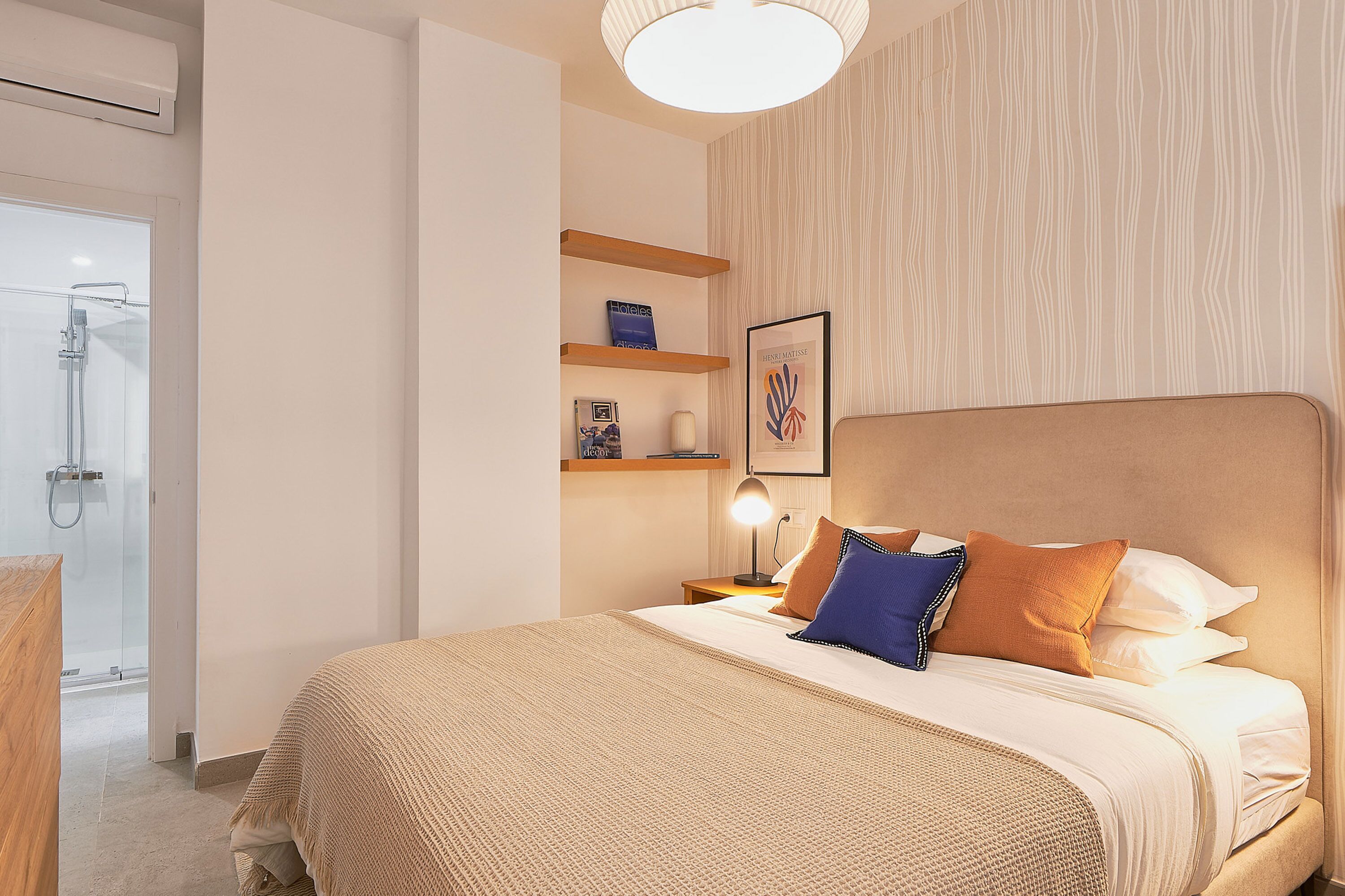 A modern bedroom with a large bed, a bedside table with a lamp, and a wall-mounted shelf with decorative items.