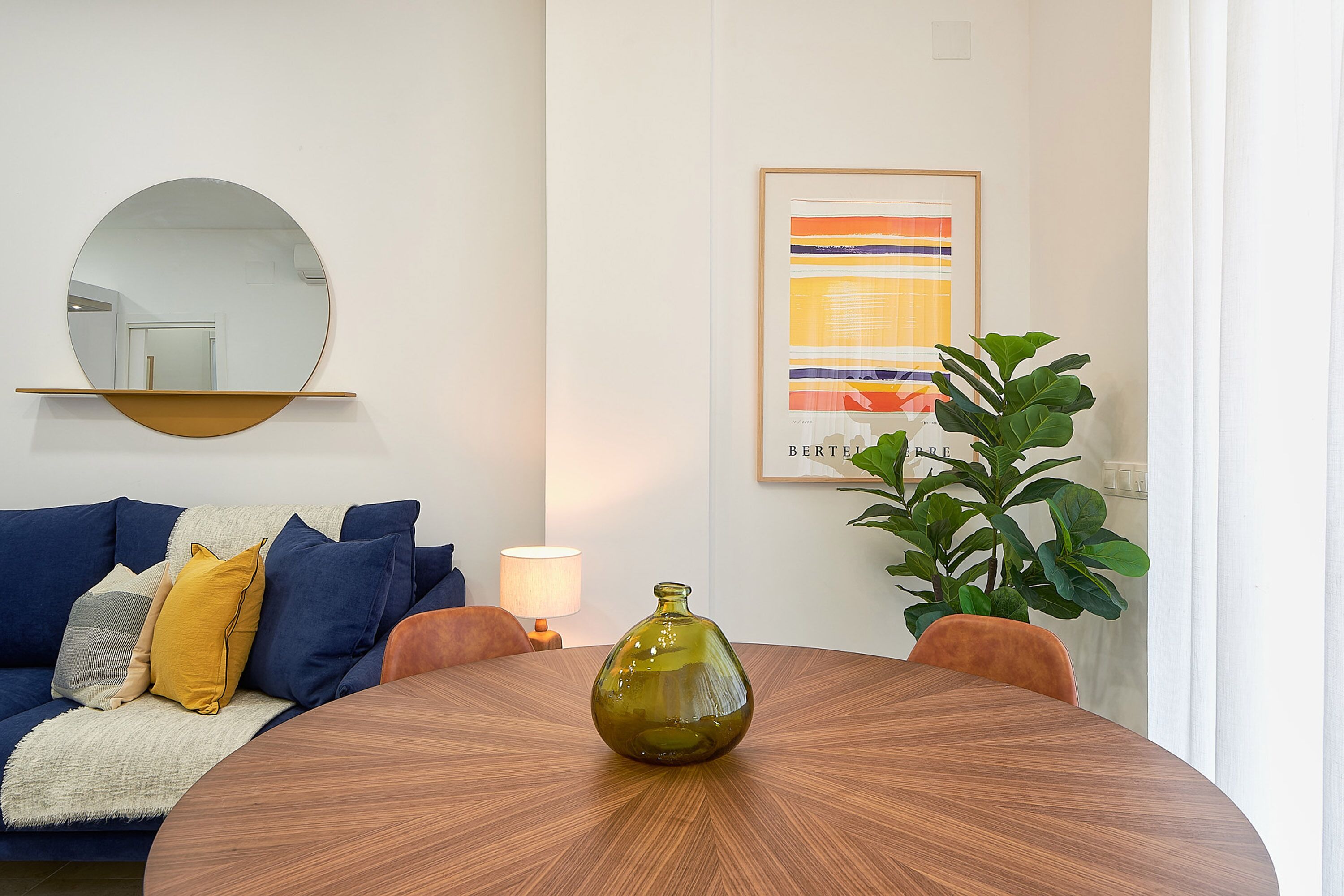 A living room with a round wooden table, a blue sofa with cushions, a mirror, a framed abstract painting, and a potted plant.