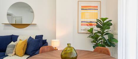 A living room with a round wooden table, a blue sofa with cushions, a mirror, a framed abstract painting, and a potted plant.