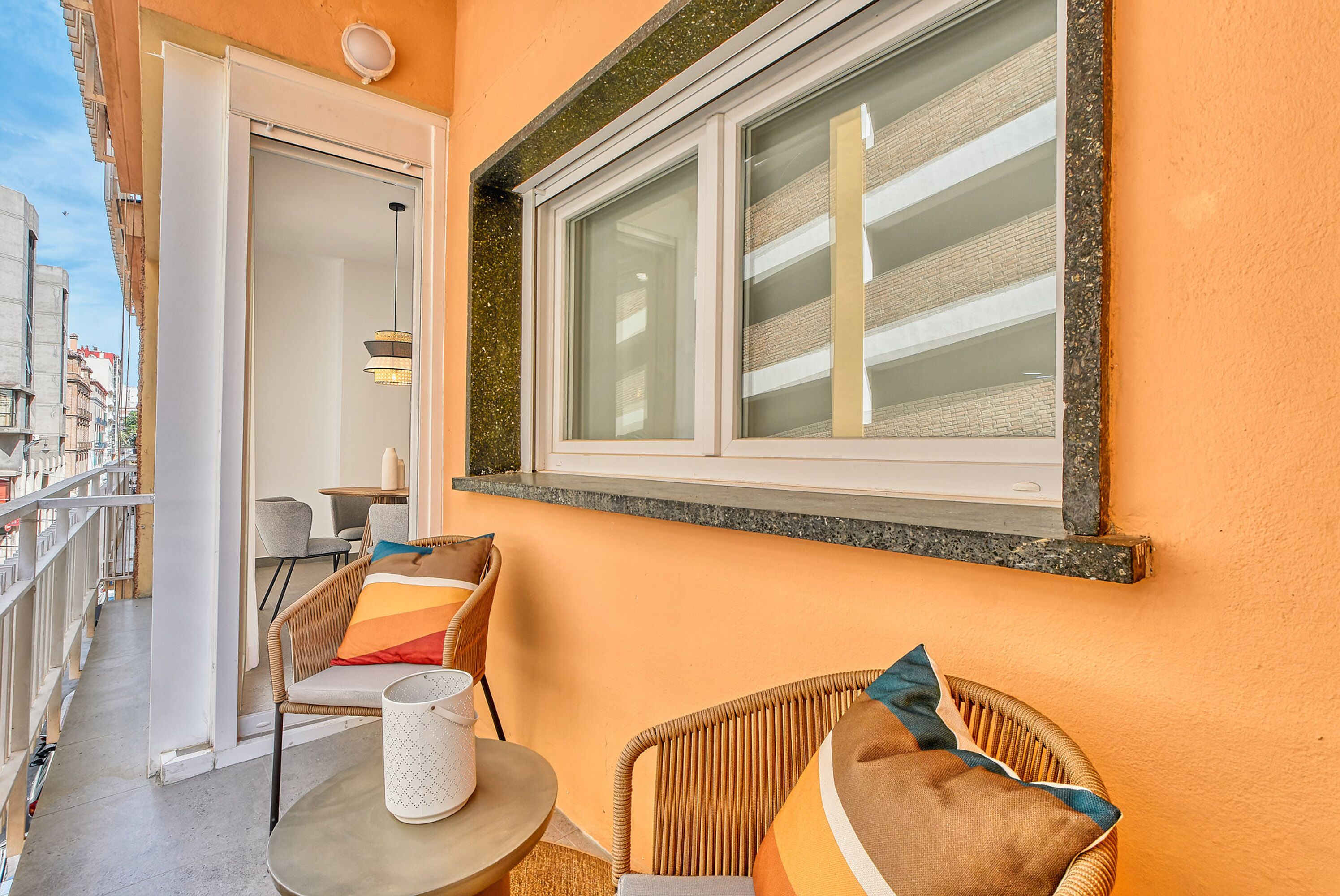 Un balcón con sillas de mimbre, una mesita y una ventana con vista a un edificio.