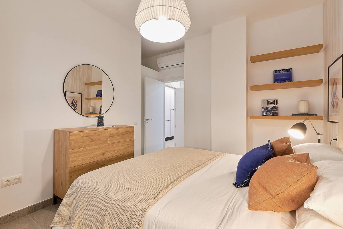 A bedroom with a bed, bedside table, mirror, and shelves with books and a lamp.