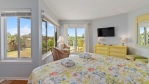 A bedroom with a large bed, a TV mounted on the wall, a chair by the window, and a yellow dresser.